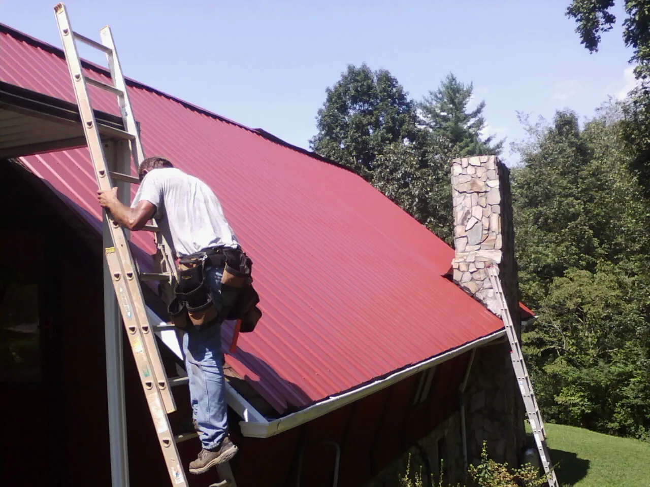 Roofer on a ladder beside a red metal roof and stone chimney — Soffit & Fascia Repair by Roofing Brothers in Galena Park